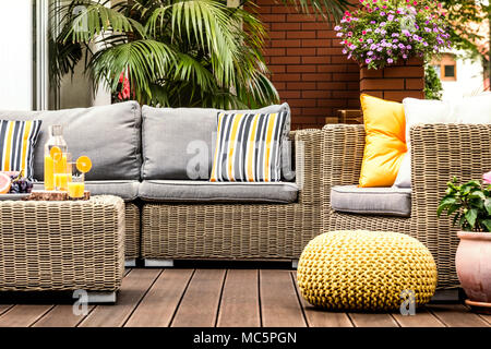 Pouf giallo accanto a una poltrona di rattan sulla terrazza in legno con cuscini a strisce sul divano Foto Stock