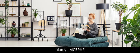 Giovane donna in bicchieri seduto su un materasso futon e la lettura di un libro in bianco sala interna con angolo studio Foto Stock