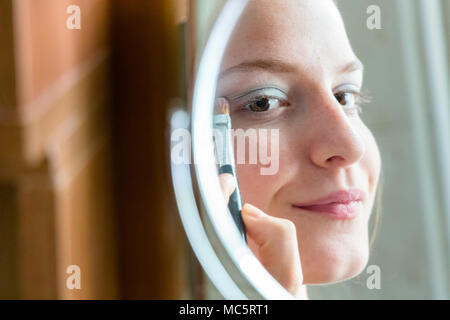 Ragazza adolescente applicando ombretto cercando la sua riflessione su uno specchio rotondo a casa Foto Stock