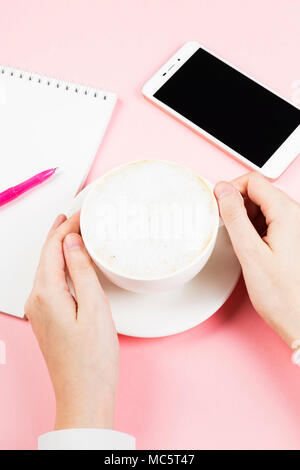 Mani femminili tenere con tazza cappuccino. Il concetto di pianificazione mattino - cappuccino, notebook, penna, telefono su sfondo rosa Foto Stock