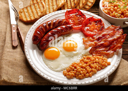 Prima colazione inglese con salsicce, pomodori grigliati, uova, pancetta, fagioli e pane sulla piastra bianca Foto Stock