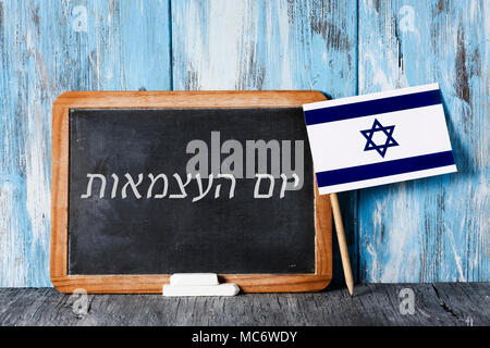 Un di legno-incorniciato lavagna con il testo il giorno di indipendenza scritta in ebraico in esso, alcuni pezzi di gesso e una bandiera israeliana su un rustico di una superficie di legno Foto Stock
