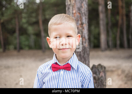 Ritratto di carino divertente piccolo ragazzo caucasico all'esterno. Positivo bionda sorridente ragazzo indossa maglietta blu e rosso il filtro bow tie guarda la fotocamera. Colore orizzontale Foto Stock