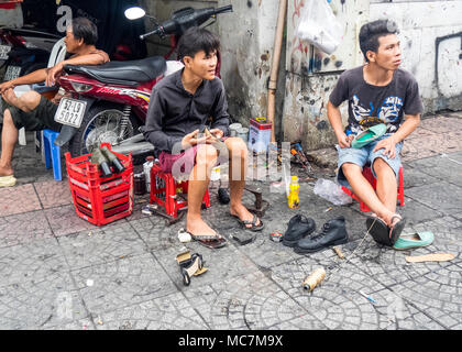 Due uomini seduti su sgabelli di plastica la riparazione di scarpe, lavorando sul marciapiede in una strada a Ho Chi Minh City, Vietnam. Foto Stock