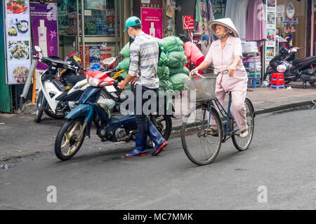 Un'anziana donna che indossa un abito rosa e un cappello conico in bicicletta su una strada nella città di Ho Chi Minh, Vietnam. Foto Stock