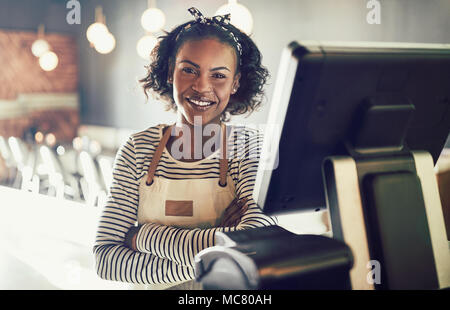 Sorridente giovane cameriera africana di indossare un grembiule in piedi da un punto terminale di vendita mentre si lavora in un ristorante alla moda Foto Stock