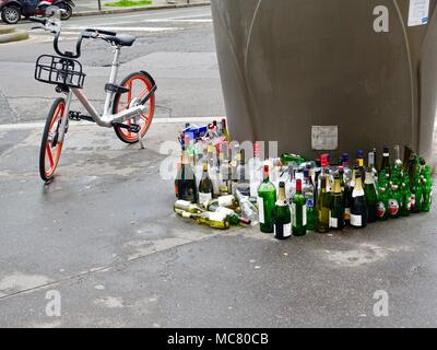 Un assortimento di bottiglie di vetro allineati ordinatamente davanti a una grande comunità, contenitore di riciclaggio sul cordolo con una bicicletta accanto ad essa. Parigi, Francia. Foto Stock