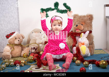 Felice adorabile bambina kidin un abito rosso e una testa di nastro, costituito di fiori è seduto sui tappeti e giocare con i giocattoli di Natale Foto Stock