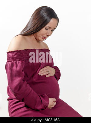 Attraente donna incinta con le mani sulla sua pancia seduto isolato su bianco Foto Stock