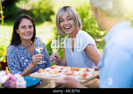 Gruppo di amici in quarantenni si sono riuniti intorno ad un tavolo in giardino per condividere un pasto. Un uomo offre spuntini a disposizione degli ospiti Foto Stock
