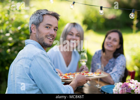 Gruppo di amici in quarantenni si sono riuniti intorno ad un tavolo in giardino per condividere un pasto. Un uomo offre spuntini per gli ospiti mentre guardando la telecamera Foto Stock