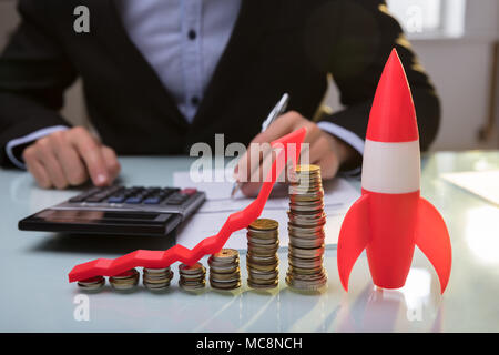 Close-up di razzo rosso oltre a monete impilate e la freccia mostra la direzione verso l'alto sulla scrivania in legno Foto Stock
