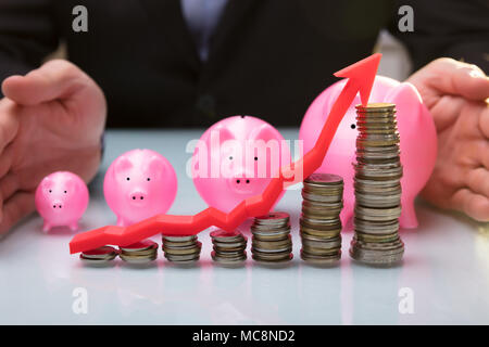 Close-up di Agente protezione rosa Salvadanaio dietro le monete impilate e la freccia mostra la direzione verso l'alto Foto Stock