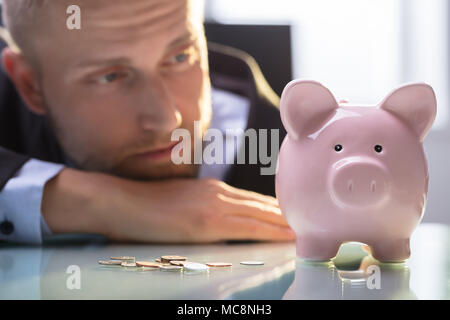 Close-up di un imprenditore depresso guardando le monete e Rosa Piggybank sulla scrivania riflettente Foto Stock