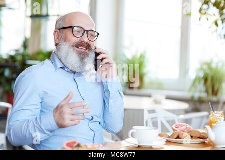 Senior uomo con barba grigia seduti a tavola in cafe e parlando su smartphone di tazza di tè Foto Stock