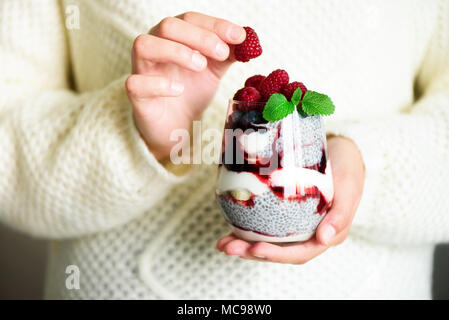 Ragazza in bianco maglione detiene il vetro con chia dessert di latte, Lamponi Mirtilli, menta. Colazione sana nozione. Spazio di copia, close up Foto Stock