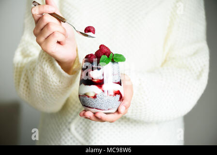 Ragazza in bianco maglione detiene il vetro con chia dessert di latte, Lamponi Mirtilli, menta. Colazione sana nozione. Spazio di copia, close up Foto Stock