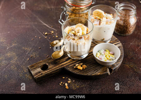 Una sana dolce fatto di yogurt e una banana, granola e i dadi su un tondo in legno tagliere su sfondo scuro Foto Stock
