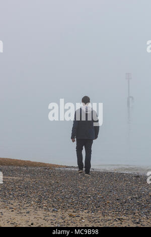 Uomo a camminare lungo una spiaggia sul suo proprio solo su di una nebbia o nebbioso giorno con ghiaia sul foreshore. Lonliness e solitudine camminare da solo vicino al mare. Foto Stock