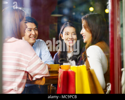 Quattro felice giovani asiatici adulti maschi e femmine rilassante conversazione in chat in coffee shop dopo lo shopping, shot attraverso il vetro di una finestra. Foto Stock