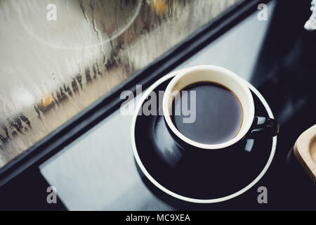 Nero caffè tazza sul tavolo nel ristorante cafe vicino alla finestra quando piove nel giardino al di fuori del negozio, il cibo e le bevande concetto,Leisure lifestyle. Foto Stock