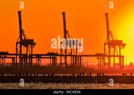 Rosso arancione bruciato il cielo al tramonto contro l'impostazione caldo sole alto contrasto pesanti carichi industriali gru da carico in Port Melbourne della Baia di Port Phillip Foto Stock