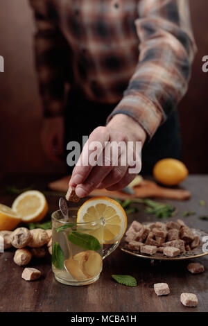 Tazza di vetro di tè allo zenzero con miele , limone e menta sul vecchio tavolo in legno. Messa a fuoco selettiva .uomo mette un pezzo di zucchero di canna in una tazza di tè. Foto Stock