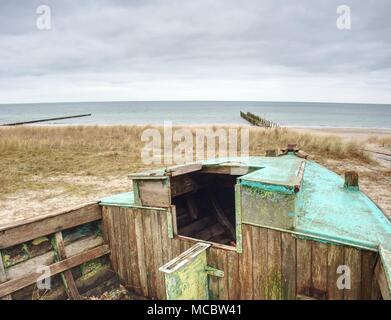 Abbandonate la barca disastrate bloccati nella sabbia. Vecchia barca in legno sulla riva sabbiosa della spiaggia. Tramonto sulla spiaggia. Vegetazione rada. Foto Stock