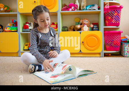 Piccola ragazza seduta sul pavimento e thumbing un libro. insegnamento in una scuola materna. o in una scuola elementare Foto Stock