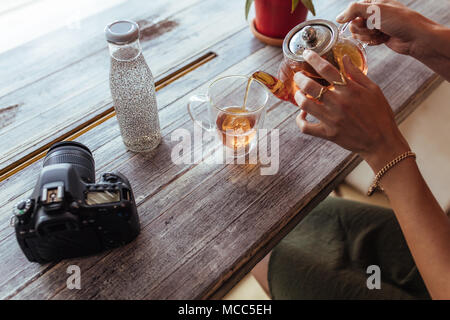 In prossimità di una donna versando il tè in una tazza da una teiera in vetro. Donna preparando il tè mentre una telecamera professionale e una bottiglia riempita con succhi di frutta sono luogo Foto Stock