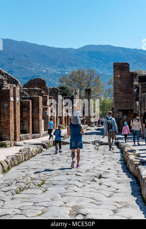 Kids in Pompei esplorare una strada rocciosa con resti di strutture e di colline in background con la famiglia, un genitore mamma trasporta un bambino piccolo sulle spalle Foto Stock