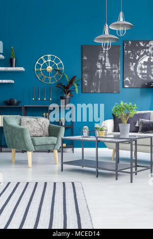 Poltrona verde accanto a un tavolo con impianto contro la parete blu con oro Orologio rotondo in living room interior Foto Stock