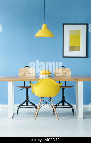 Stile minimal interni simmetrica con una luce gialla appesa sopra un tavolo di legno con un melone in un piatto da portata, sedie e un poster su una parete blu Foto Stock