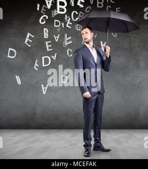 Imprenditore si erge con ombrello sotto la caduta di lettere oltre il muro grigio Sfondo. Foto Stock