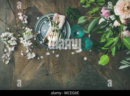 Flat-lay di primavera vacanze di Pasqua messa in tavola. Gara di mandorle fiori fioriscono sui rami e le peonie, piatti, bicchieri e posate su woode vintage Foto Stock