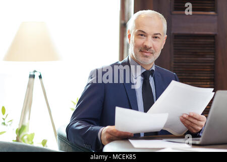 Ritratto di fiducioso grey-haired uomo caucasico in abbigliamento business seduta a tavola con documenti e sorridente in telecamera Foto Stock