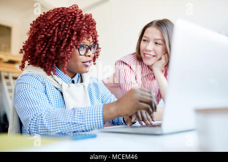 Elegante americano africano donna con capelli rossi lavora sul computer portatile con caucasica collega femmina e sorridente Foto Stock