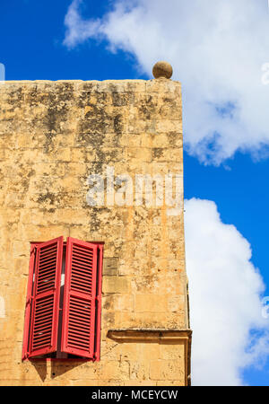 Mdina, Malta isola. Persiane rosse finestra su una pietra arenaria gialla nel muro della vecchia città medievale. Cielo blu sullo sfondo Foto Stock