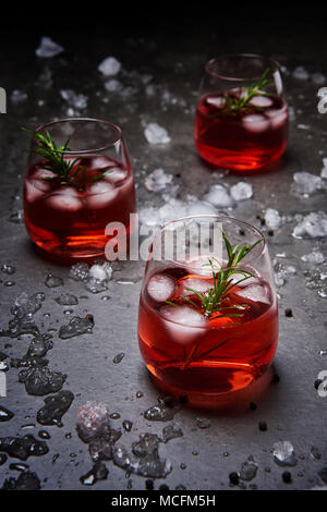 Melograno cocktail con ghiaccio e rosmarino circondato con ghiaccio tritato sul calcestruzzo nero lo sfondo. Foto Stock