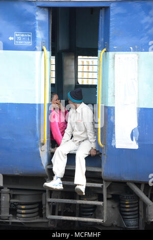 Uomo che fa una donna ridere mentre il pendolarismo su un treno indiano, seduta precariamente nella porta aperta Foto Stock