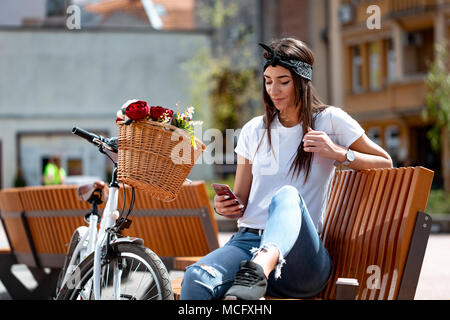 Sorridente giovane donna scrivere messaggi su smartphone e seduta su una panchina in una giornata di sole, accanto alla moto con cesto fiorito. Foto Stock