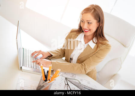 Sorridente giovane donna architetto operante su laptop alla scrivania in ufficio. Foto Stock