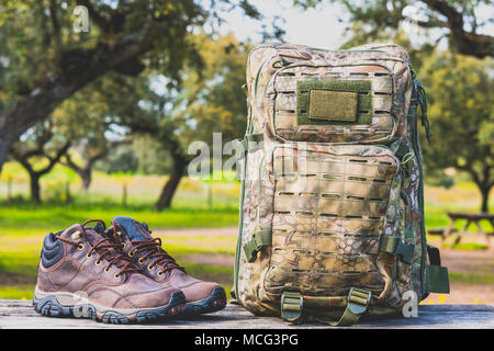 Scarpe da trekking e zaino camouflage su una tavola di legno in natura Foto Stock