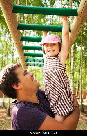 Padre aiutare la propria figlia appesa sulla Monkey bar in un parco giochi, sorridente e trascorrere del tempo insieme. Foto Stock