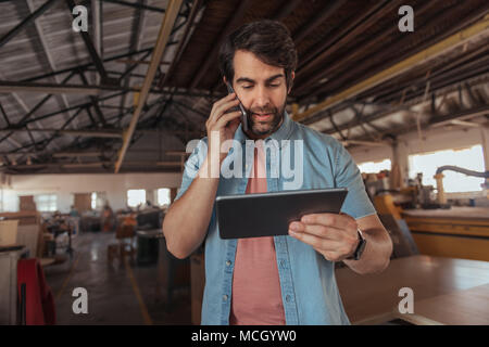 Giovane falegname con la barba a parlare su un cellulare e utilizzando una tavoletta digitale mentre si lavora nel suo grande studio di macchine per la lavorazione del legno Foto Stock