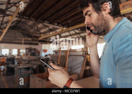 Giovane falegname con la barba a lavorare da solo nel suo grande negozio di macchine per la lavorazione del legno utilizzando una tavoletta digitale e parlando al cellulare Foto Stock