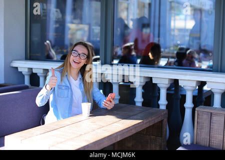 Ragazza che mostra i pollici fino a street cafe e mantenendo lo smartphone. Foto Stock