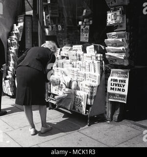 Londra, 1967, una signora si piega giù per leggere i titoli dei giornali internazionali, nazionali e britannici locali come il posto di Kensington, su un newstand fuori un chiosco del tabacco del marciapiede. In mostra presso lo stand vi sono i fortunati medaglioni 'World Cup Willie', souvenir per il torneo di calcio della Coppa del mondo 1966 che si è tenuto in Gran Bretagna, con la finale al Wembley Stadium. Foto Stock