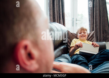 Little Boy e suo padre stanno parlando mentre sdraiato sul divano insieme. Il ragazzino è in possesso di una tavoletta digitale. Foto Stock