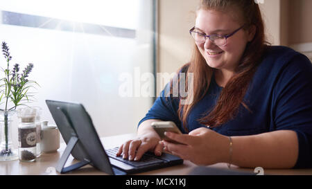 Caucasian ragazza sorridente mentre guardando il suo cellulare durante la sua tazza di mattina di cappachino mentre lei controlla la sua posta su una tavoletta elettronica nel suo bar preferito con una luce brillante. Foto Stock